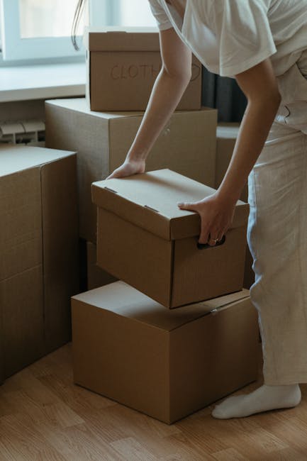 A person wearing a white shirt and light-colored trousers is in an indoor space, surrounded by multiple cardboard moving boxes of various sizes, some labeled with handwritten markings such as 'CLOTH'.