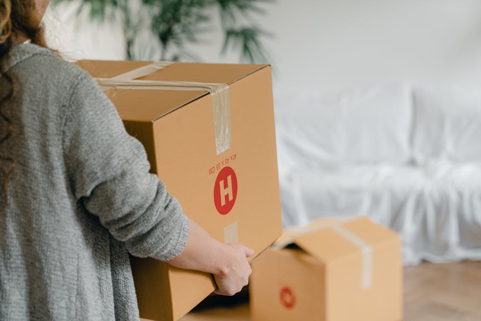 A person wearing a white shirt and light-colored trousers is in an indoor space, surrounded by multiple cardboard moving boxes of various sizes, some labeled with handwritten markings such as 'CLOTH'.