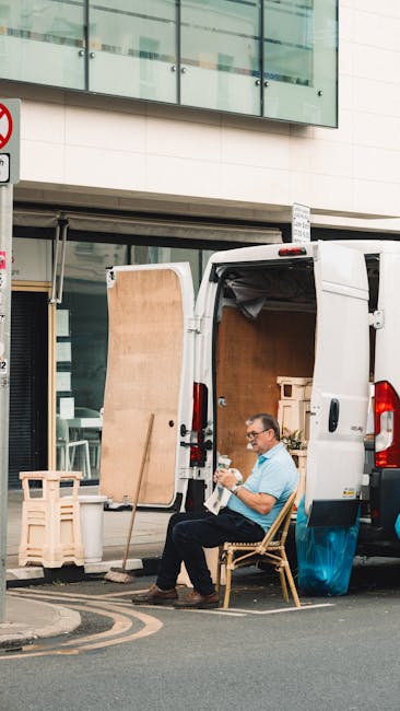 A man wearing glasses and a light blue shirt is seated outside a white moving van with its rear doors open on a city street, taking a break during a house or office relocation process. The van is park